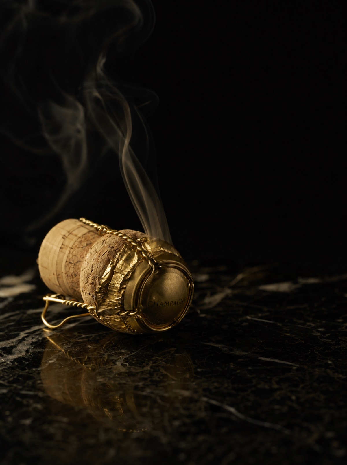 A champagne cork with gold foil on black marble, a wisp of smoke